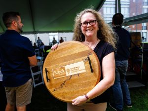 Barbara Tatarnic holds a large wooden display made from a wine barrel head featuring the Brock University Cool Climate Oenology and Viticulture Institute logo while standing under a tent at an outdoor event.