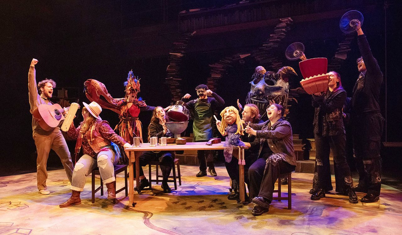 The cast of a play performs on a theatre stage, many using puppets in their performance.