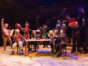 The cast of a play performs on a theatre stage, many using puppets in their performance.