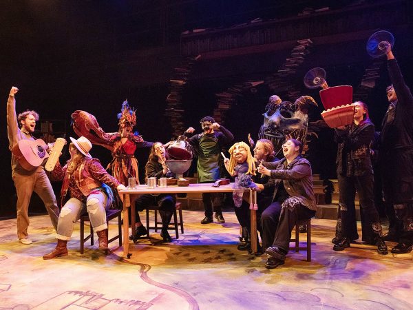 The cast of a play performs on a theatre stage, many using puppets in their performance.