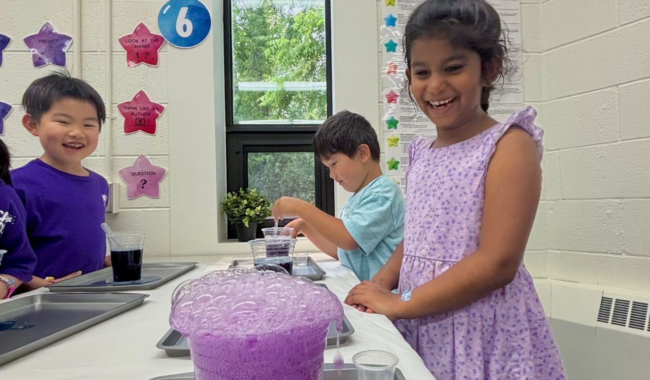 Three young children participate in a science experiment watching a bubbling purple mixture spill out of a clear container.
