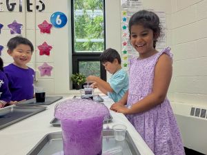 Three young children participate in a science experiment watching a bubbling purple mixture spill out of a clear container.
