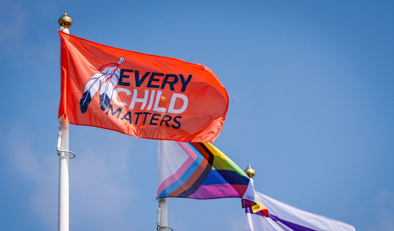 Three flag poles set against a blue sky, waving in the wind is an Every Child Matters flag in the forefront, a Progress Pride flag in the middle, and a Two Row Wampum flag in the background.