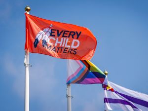 Three flag poles set against a blue sky, waving in the wind is an Every Child Matters flag in the forefront, a Progress Pride flag in the middle, and a Two Row Wampum flag in the background.