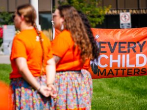 Three women wearing orange shirts and floral ribbon skirts stand outdoors, facing away from the camera, with an Every Child Matters flag hanging low on a flagpole behind them.