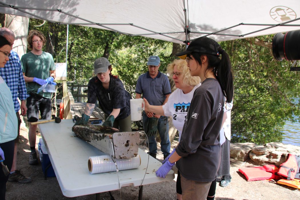 A group of people are gathered around a table looking at a steel rod covered with mud. One woman is leaning over and touching the rod, while another woman i9s holding a jug over the rod.