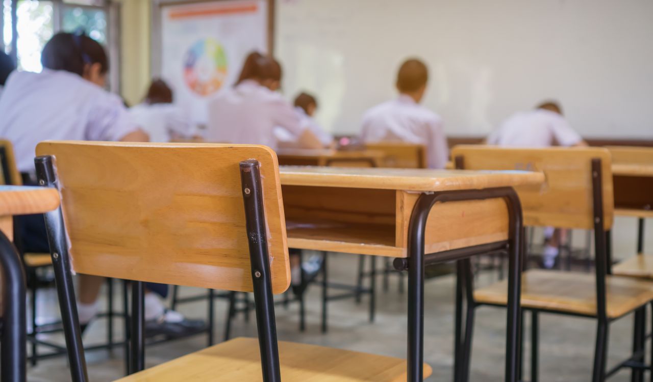 An empty chair in a classroom with students seated at desks in the background.