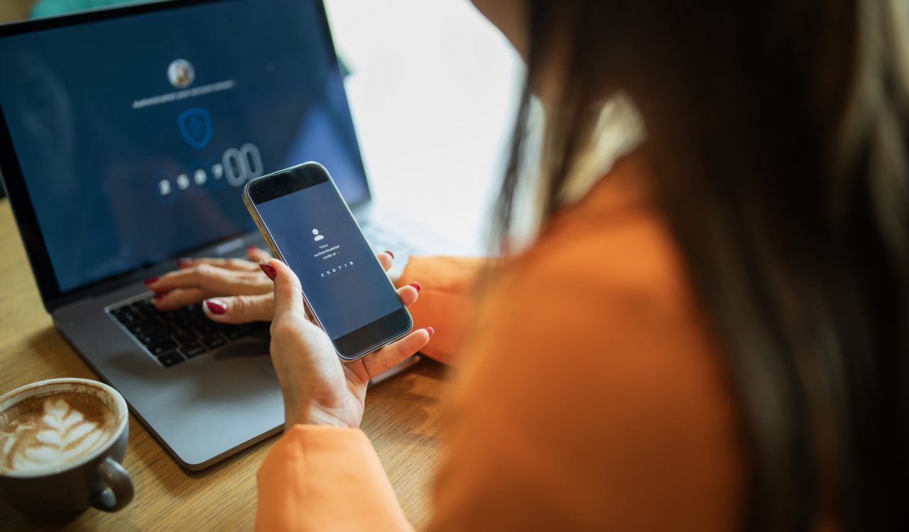 Close-up of a person holding a phone with a two-factor authentication on the screen. The person is using a laptop with the other hand.