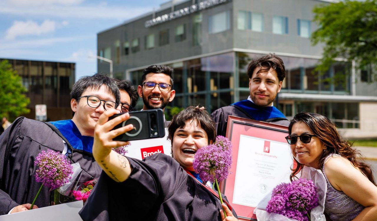 A group of six people smile as they pose for a selfie outside of Brock University’s Goodman School of Business. The person in front holds up a cellphone to take the photo. Three of the people are wearing graduation robes and holding diplomas.