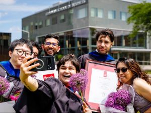 A group of six people smile as they pose for a selfie outside of Brock University’s Goodman School of Business. The person in front holds up a cellphone to take the photo. Three of the people are wearing graduation robes and holding diplomas.