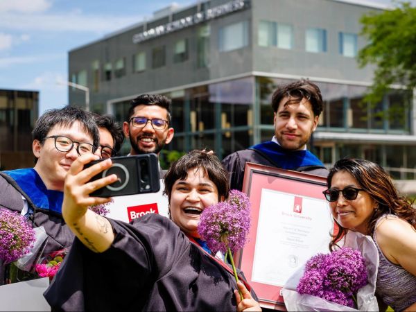 A group of six people smile as they pose for a selfie outside of Brock University’s Goodman School of Business. The person in front holds up a cellphone to take the photo. Three of the people are wearing graduation robes and holding diplomas.