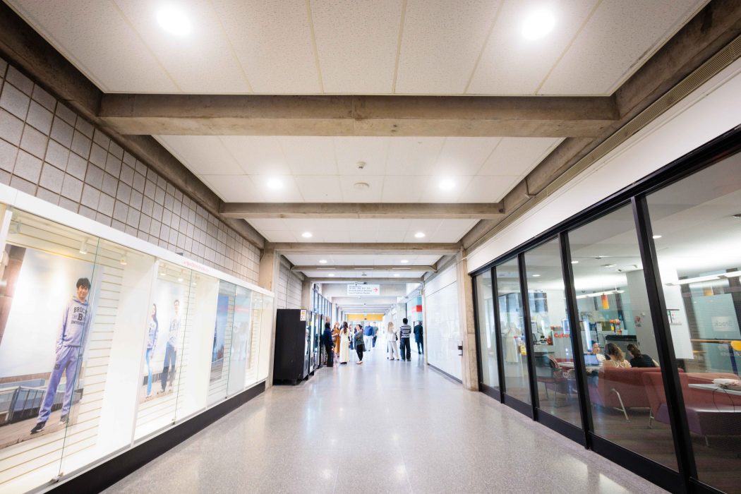 A bright indoor corridor on the Brock University campus features exposed concrete beams, polished floors and glass-fronted spaces on both sides. Display cases line the left wall, while people gather and sit in lounge areas down the right side of the hallway.