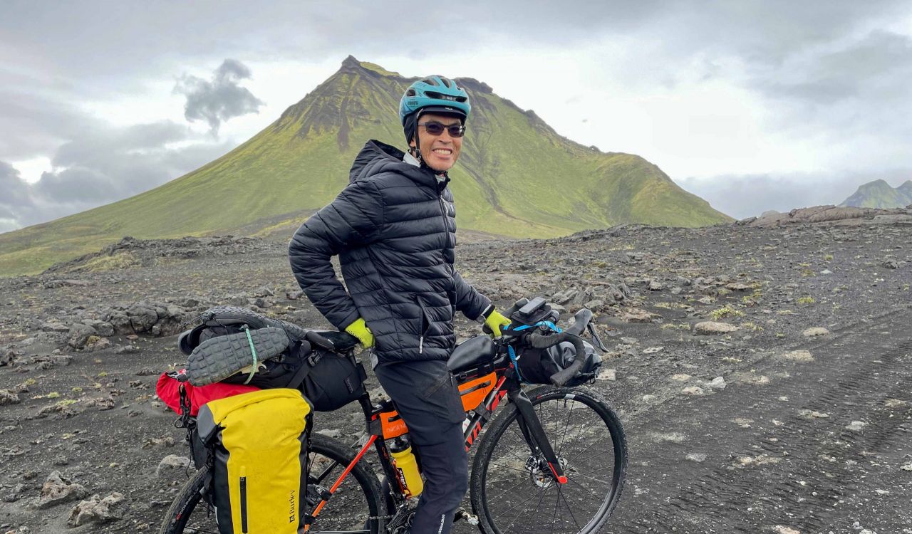 A man rests with his bicycle on a patch of flat ground with a volcano in the background.