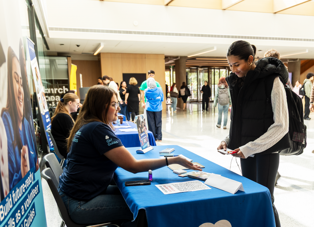 A woman stands in front of an information table in the Rankin Family Pavilion and listens as another woman, seated behind the table, speaks.