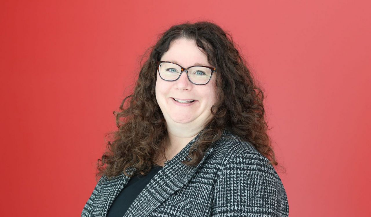Head-and-shoulders portrait of Jennifer Guarasci with long curly dark hair and glasses wearing a black top and grey patterned blazer, standing against a solid red background.