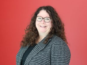 Head-and-shoulders portrait of Jennifer Guarasci with long curly dark hair and glasses wearing a black top and grey patterned blazer, standing against a solid red background.