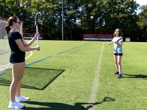 Two women dressed in athletic clothes stand facing each other, a short distance apart, on an outdoor athletic field. They are holding lacrosse sticks, tossing a ball between them.