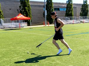 A man in black athleticwear chases a ball on an outdoor athletic field, holding a lacrosse stick.