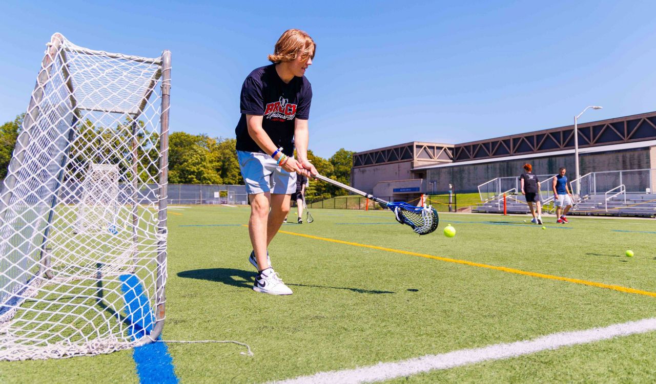A man holds a lacrosse stick with both hands as he stands on an outdoor athletic field, beside a goal net.