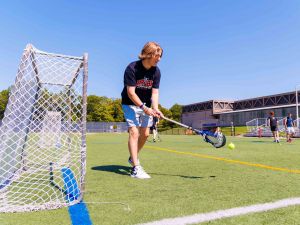 A man holds a lacrosse stick with both hands as he stands on an outdoor athletic field, beside a goal net.