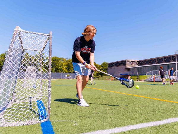 A man holds a lacrosse stick with both hands as he stands on an outdoor athletic field, beside a goal net.