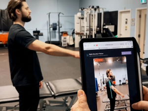 A man performs shoulder exercises while another person, holding a tablet, evaluates him.