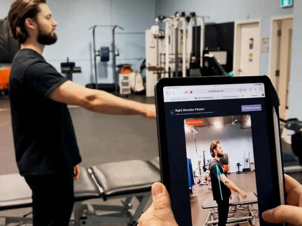A man performs shoulder exercises while another person, holding a tablet, evaluates him.