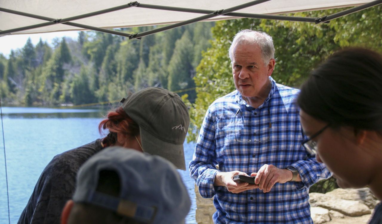 A man in a checkered shirt stands in front of three people bending over a table, with trees and water in the background.
