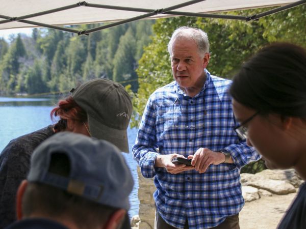 A man in a checkered shirt stands in front of three people bending over a table, with trees and water in the background.