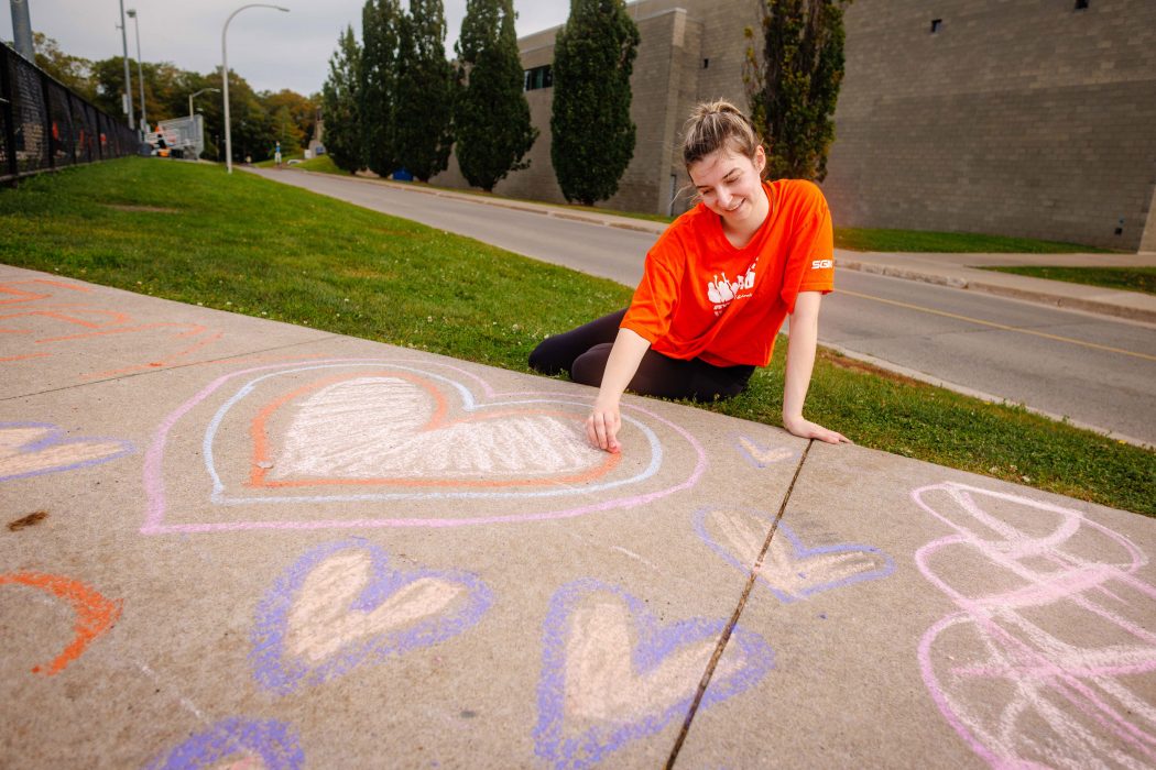 Master’s of Critical Sociology student Ally Negus BA’25 kneels on the pavement near Alumni Field, writing one of the Truth and Reconciliation Commission of Canada’s 94 Calls to Action in colourful chalk during Brock’s Chalk the Campus event.