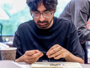 A man in a black shirt, wearing glasses, sits at a table with a window behind him. His hands are on the table, and he stares down at a beading project he is holding.