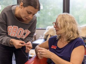 A woman, standing in an office, leans down with a beading project in her hands, showing it to a woman sitting in a chair beside her.