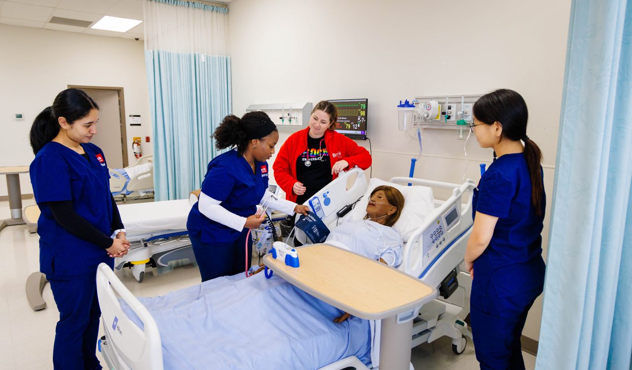 Nursing students in scrubs stand over a simulation mannequin in a hospital bed. One student is checking the blood pressure of the mannequin.