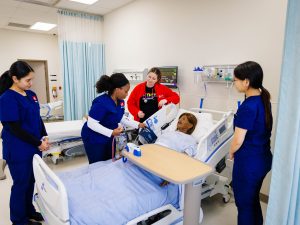 Nursing students in scrubs stand over a simulation mannequin in a hospital bed. One student is checking the blood pressure of the mannequin.