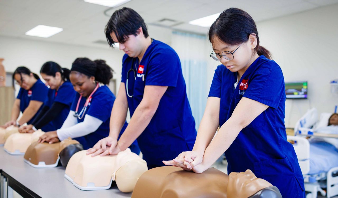 Nursing students in blue scrubs perform CPR on training manikins.