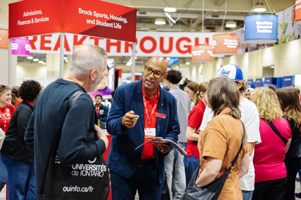 Four people stand speaking to one another in a busy convention centre space filled with Brock University branding.