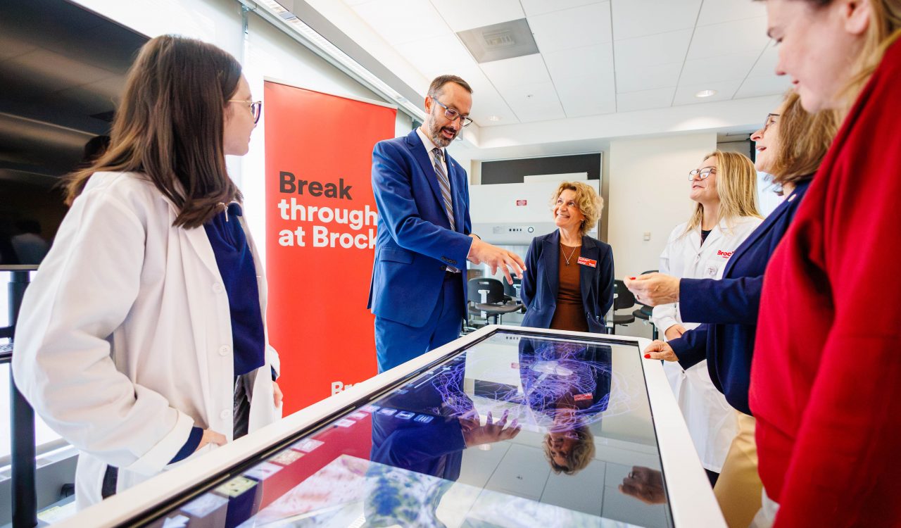 A group looks over a digital glass table featuring human anatomy.