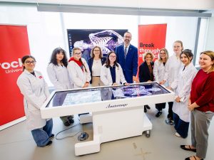 A group of people in lab coats stand behind a glass table with a rendering of a human skeleton on the screen.