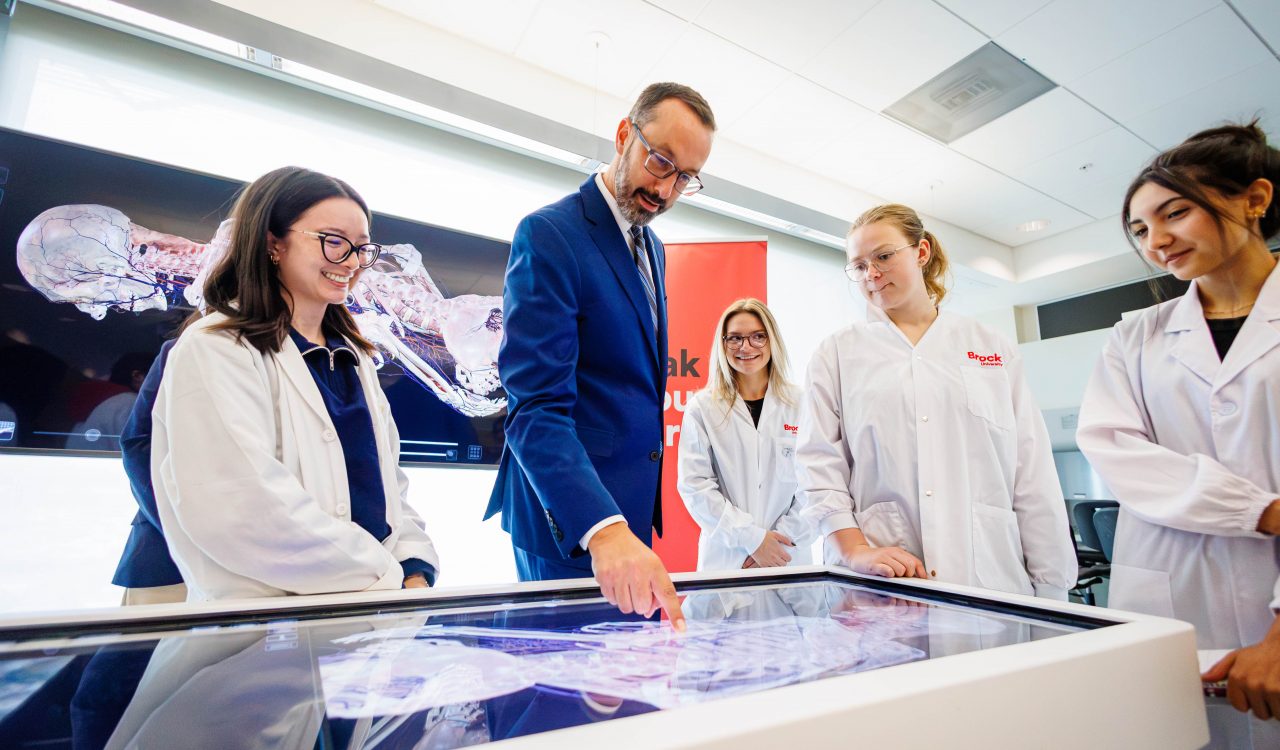 A man in a blue suit examines a digital anatomy display alongside three university students in lab coats.