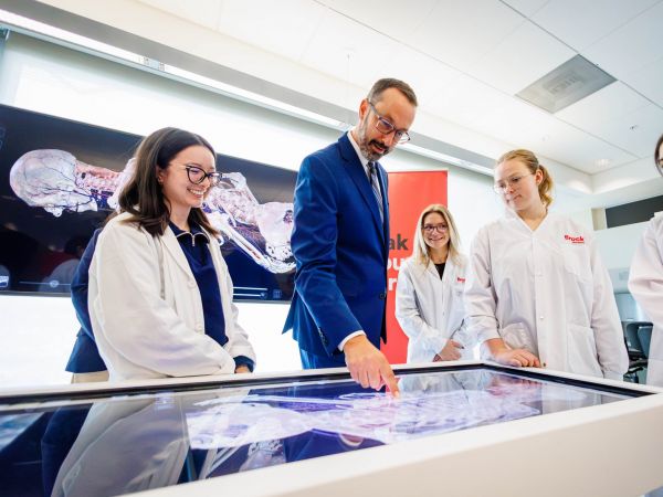 A man in a blue suit examines a digital anatomy display alongside three university students in lab coats.