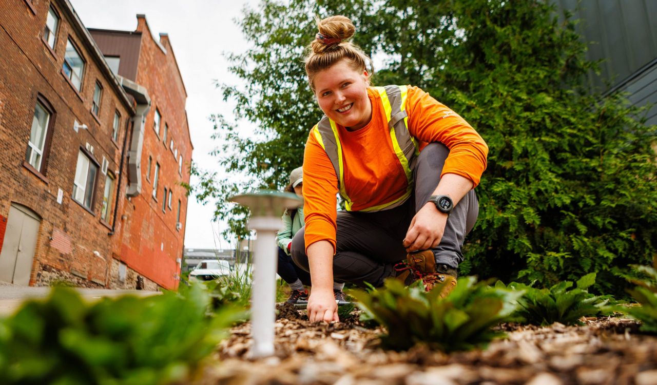 A woman kneels on the ground in a garden.