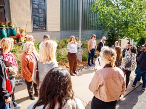 People stand in a circle listening to a woman speak at a tour of a rain garden.