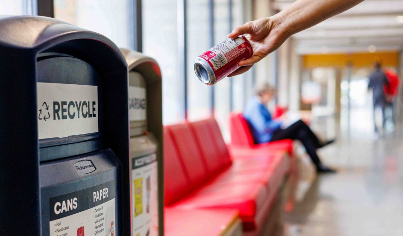 A hand holds an empty beverage can above a recycling bin in an indoor space. Red bench seating lines the background, with a seated person and another person walking in the distance.