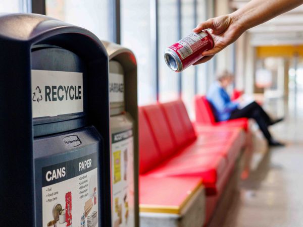 A hand holds an empty beverage can above a recycling bin in an indoor space. Red bench seating lines the background, with a seated person and another person walking in the distance.
