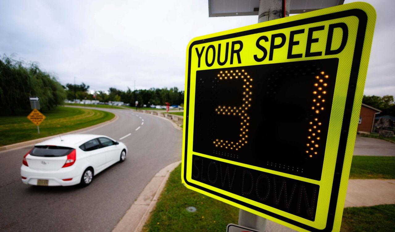 A white car drives along a curved road past a digital speed display sign reading the number 31, with grassy areas and trees in the background.