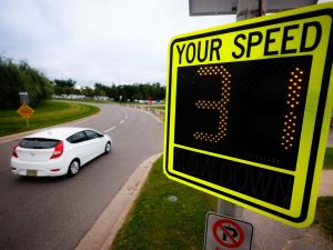 A white car drives along a curved road past a digital speed display sign reading the number 31, with grassy areas and trees in the background.