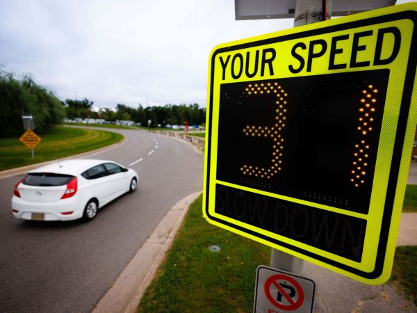 A white car drives along a curved road past a digital speed display sign reading the number 31, with grassy areas and trees in the background.