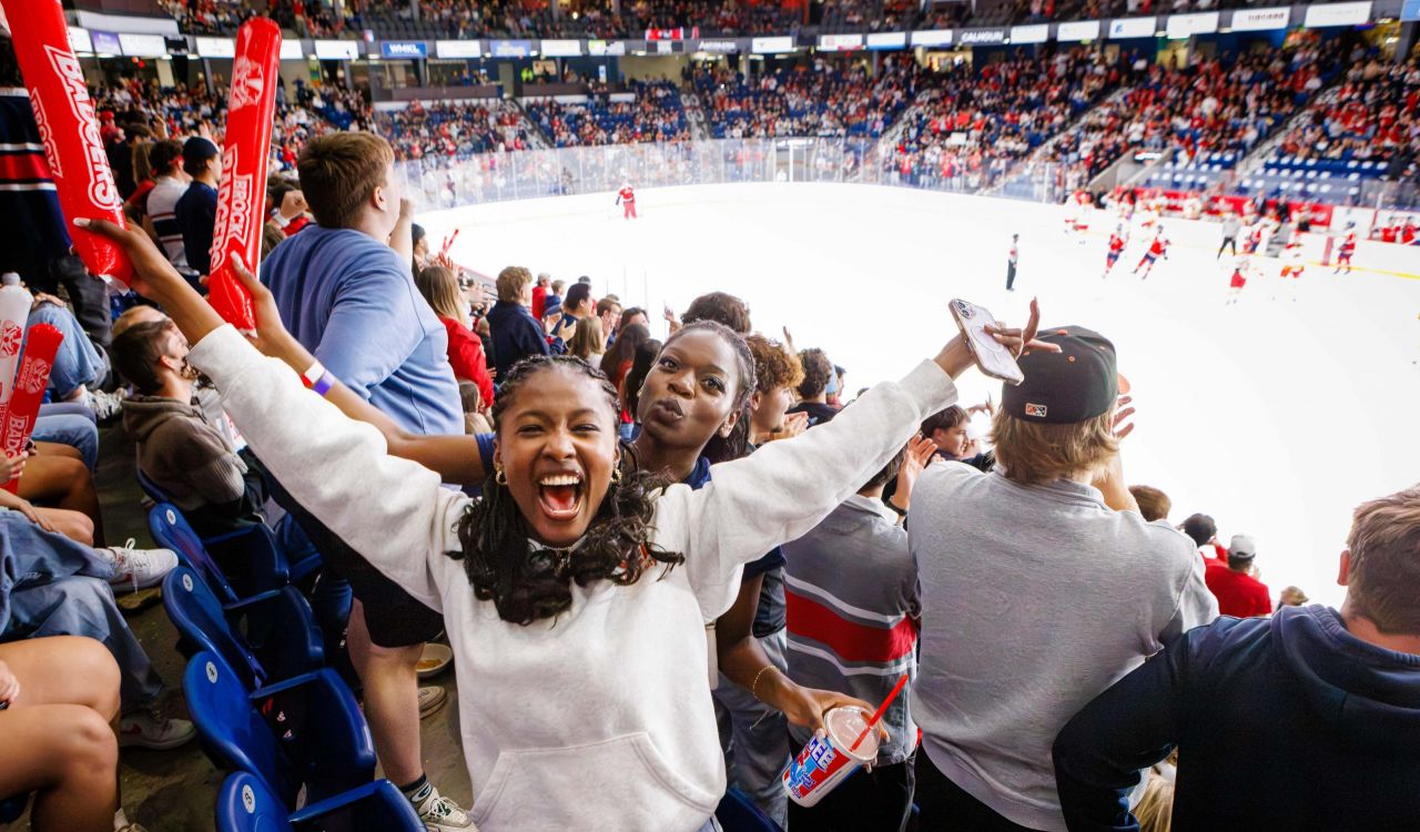 Brock University fans scream in the stands while a hockey game goes on in a packed arena.
