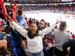 Brock University fans scream in the stands while a hockey game goes on in a packed arena.