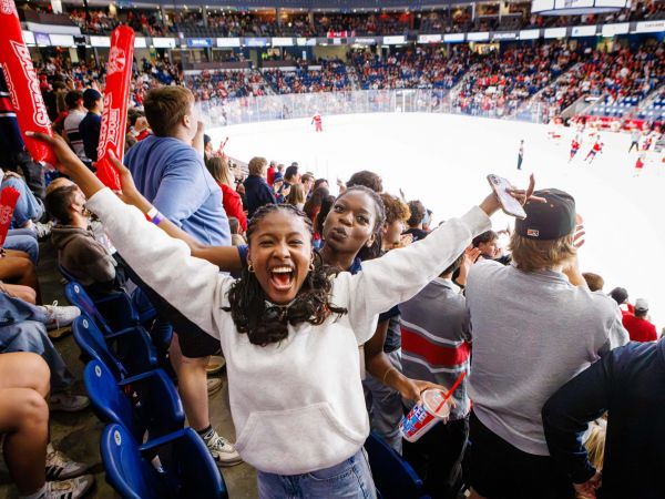Brock University fans scream in the stands while a hockey game goes on in a packed arena.